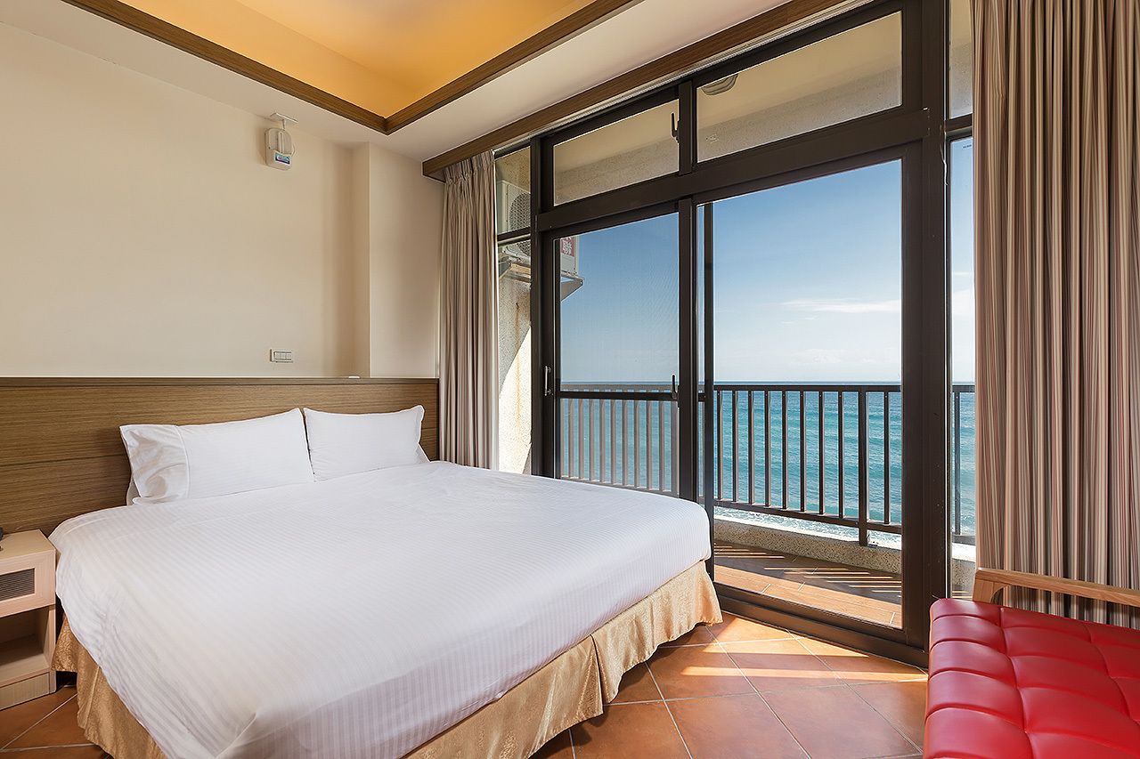 A hotel room with a large bed, a nightstand, a red chair, and a balcony with a view of the sea.