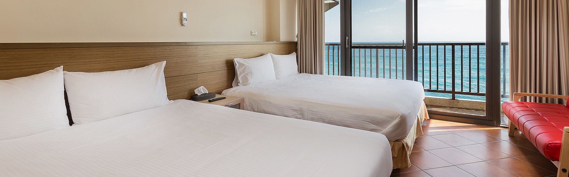 A hotel room with two beds, a red sofa, and a balcony with a view of the sea.