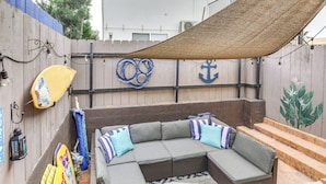 Un espace extérieur aménagé, abrité, avec un canapé d’angle, des coussins et une planche de surf.