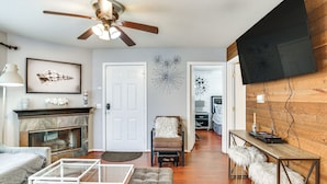 Un salon moderne doté d’une cheminée, d’un ventilateur de plafond, d’un téléviseur à écran plat fixé à un mur en bois et d’un coin repas avec des tabourets.