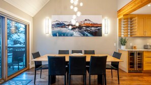 Un coin repas avec une grande table en bois et des chaises noires, un tableau représentant un paysage de montagne, et une cuisine dotée de meubles en bois et d’un réfrigérateur en acier inoxydable.