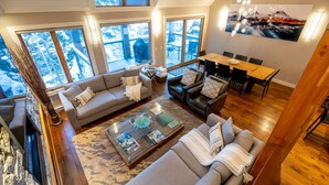 A spacious living area with a large sofa, a coffee table, and a dining table. There are multiple windows providing views of a snowy landscape.