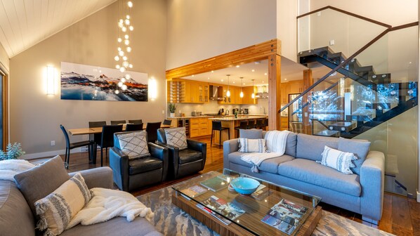 Un salon moderne comprenant un canapé, des fauteuils, une table basse avec des magazines, un coin repas avec une table et des chaises, une cuisine avec des meubles en bois et un escalier doté d’une rampe en verre.