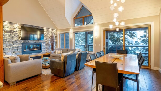 A spacious living area with a stone fireplace, comfortable seating, a dining table, and large windows offering a view of snow-covered trees.