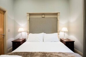 Una habitación de hotel con una cama, dos mesitas de noche con lámparas, una puerta de madera y una pared de color claro.