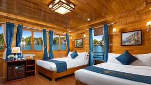 A hotel room with wooden walls and floors, two beds with blue bedding, a desk with a lamp, and a view of a body of water. - La Paci Cruises (Hai Phong)
