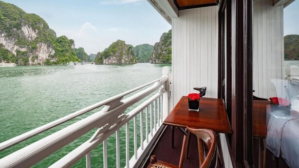 A balcony with a wooden table and chairs, overlooking a river with cliffs. - La Paci Cruises (Hai Phong)