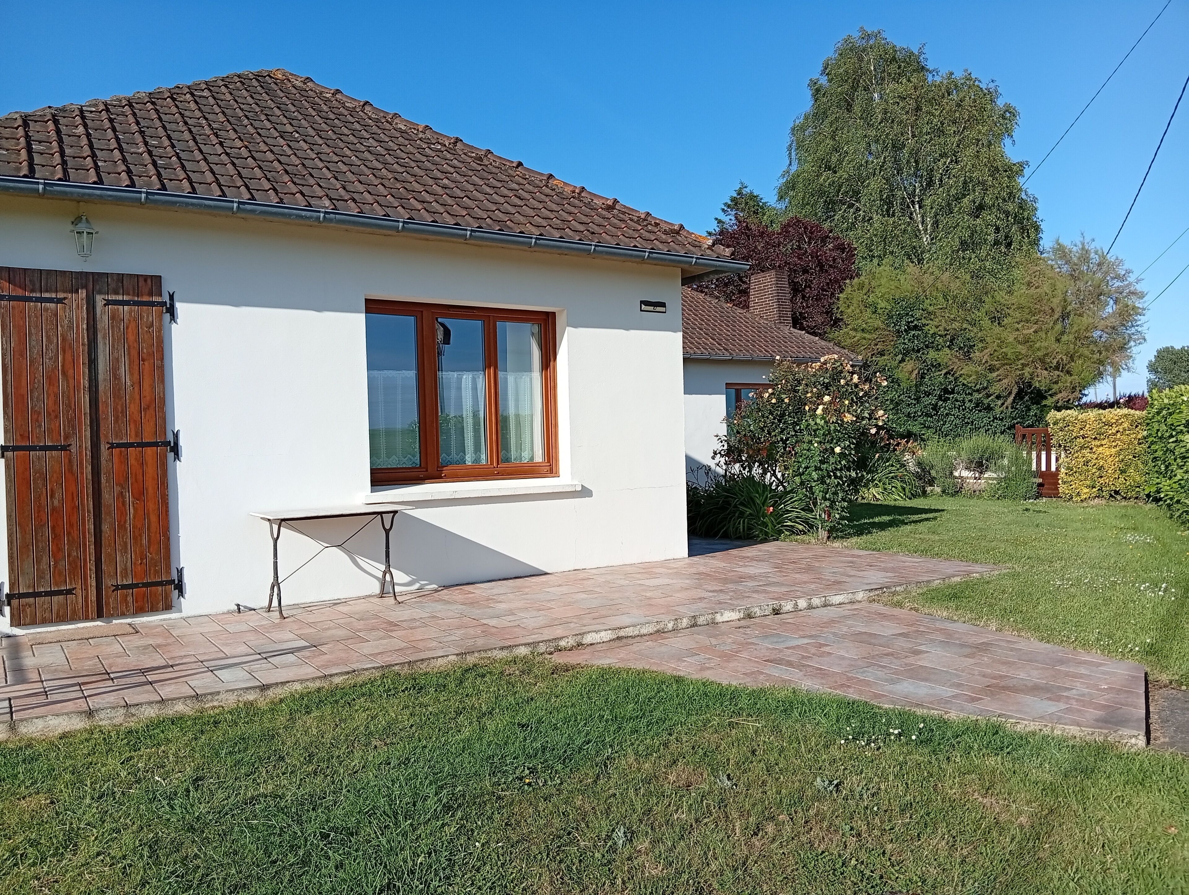 Une maison de plain-pied avec un toit en tuiles, une fenêtre avec des volets en bois et un petit espace pavé devant.