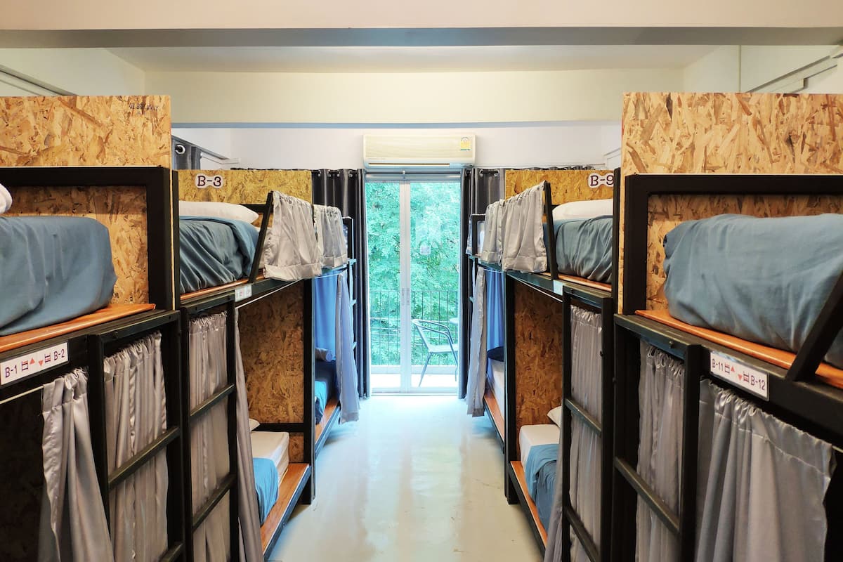 A corridor with bunk beds labeled B-1 to B-12, wooden walls, and a view of greenery through a door.