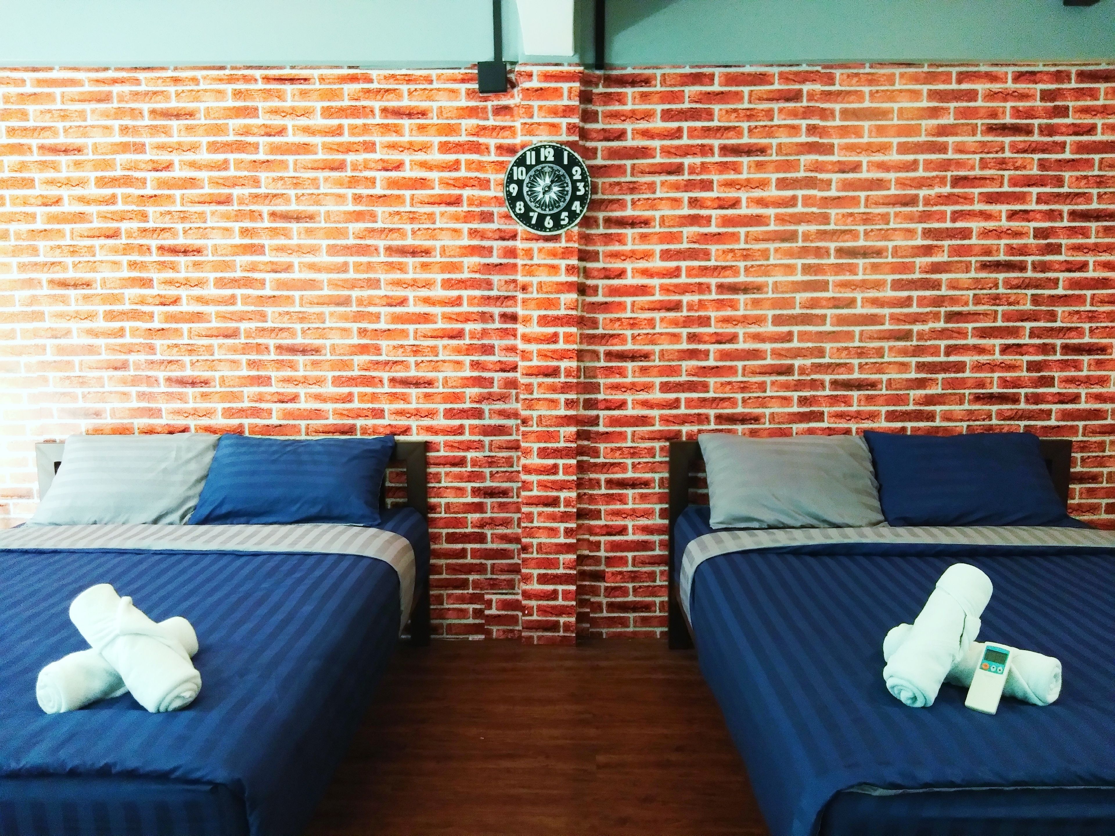 A room with two beds, each with blue bedding and pillows, against a brick wall. A round clock is mounted on the wall.