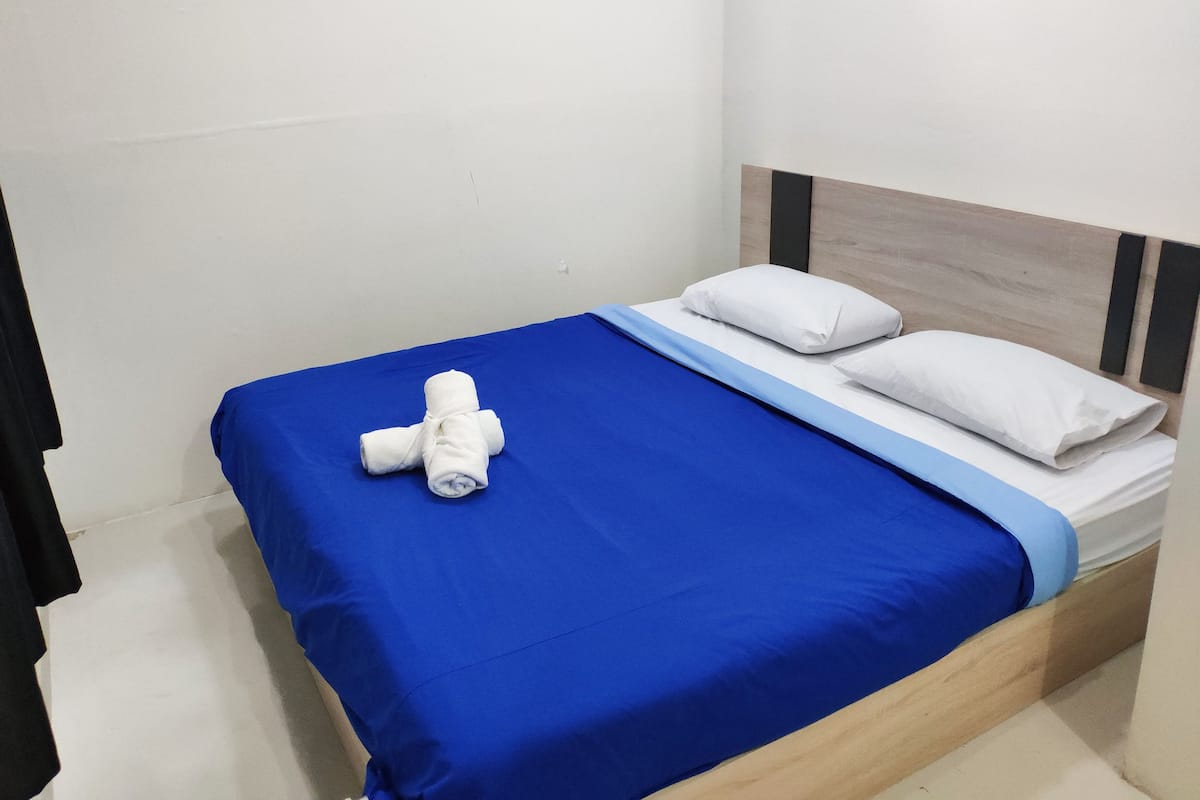 A neatly made bed with a blue bedspread and white pillows, a rolled white towel on the left side, and a wooden headboard.