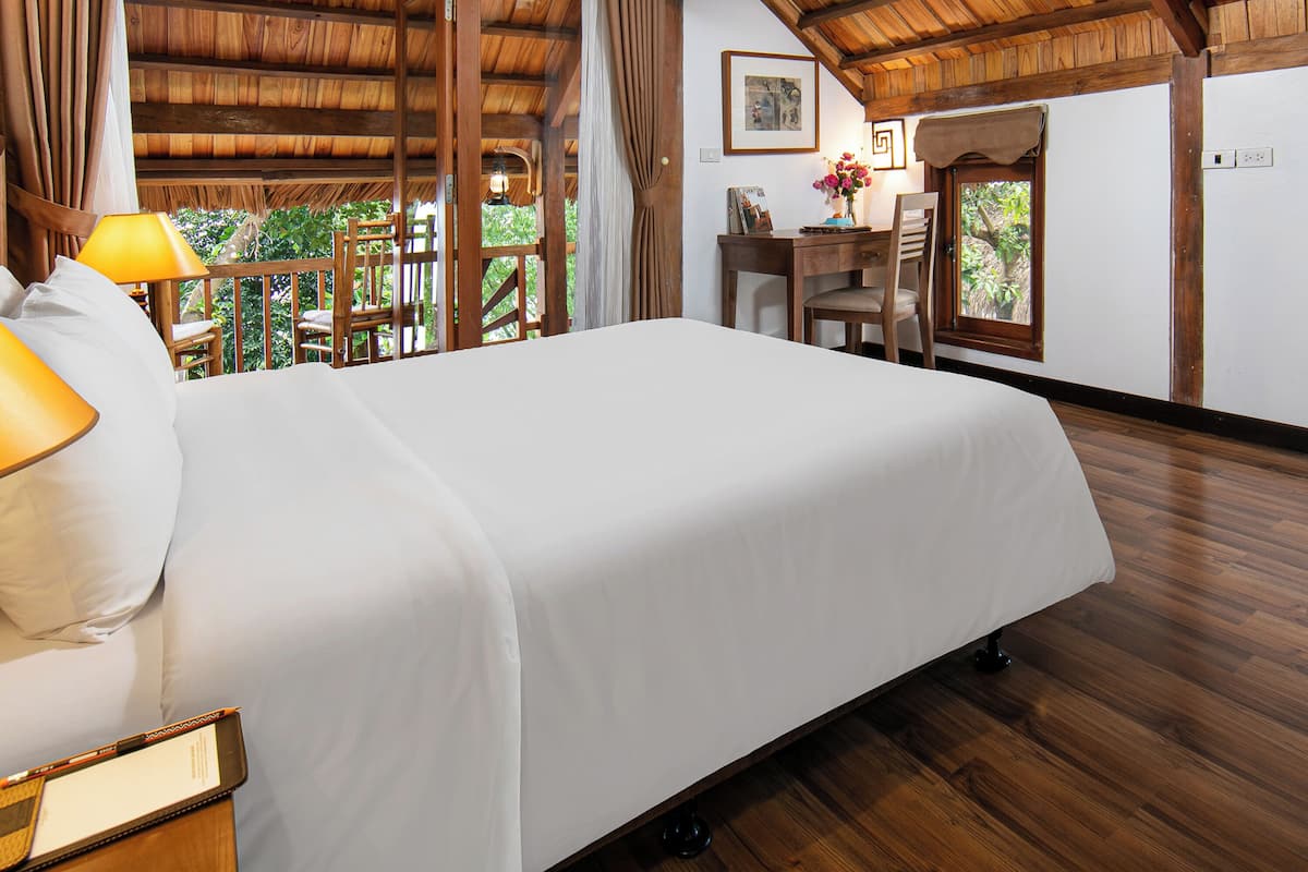 Une chambre avec un grand lit, des poutres apparentes en bois au plafond, un bureau avec une chaise et une fenêtre donnant sur un espace verdoyant.