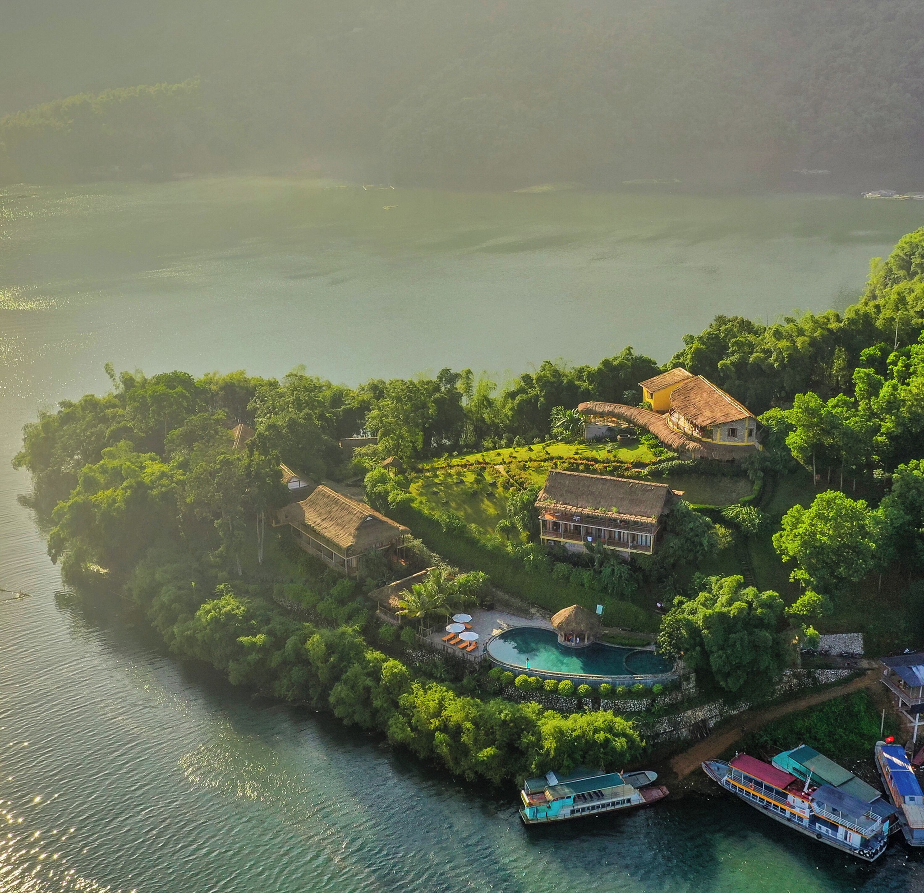 Vista aérea de una isla tropical con una casa grande, una piscina y barcos amarrados cerca de la costa.