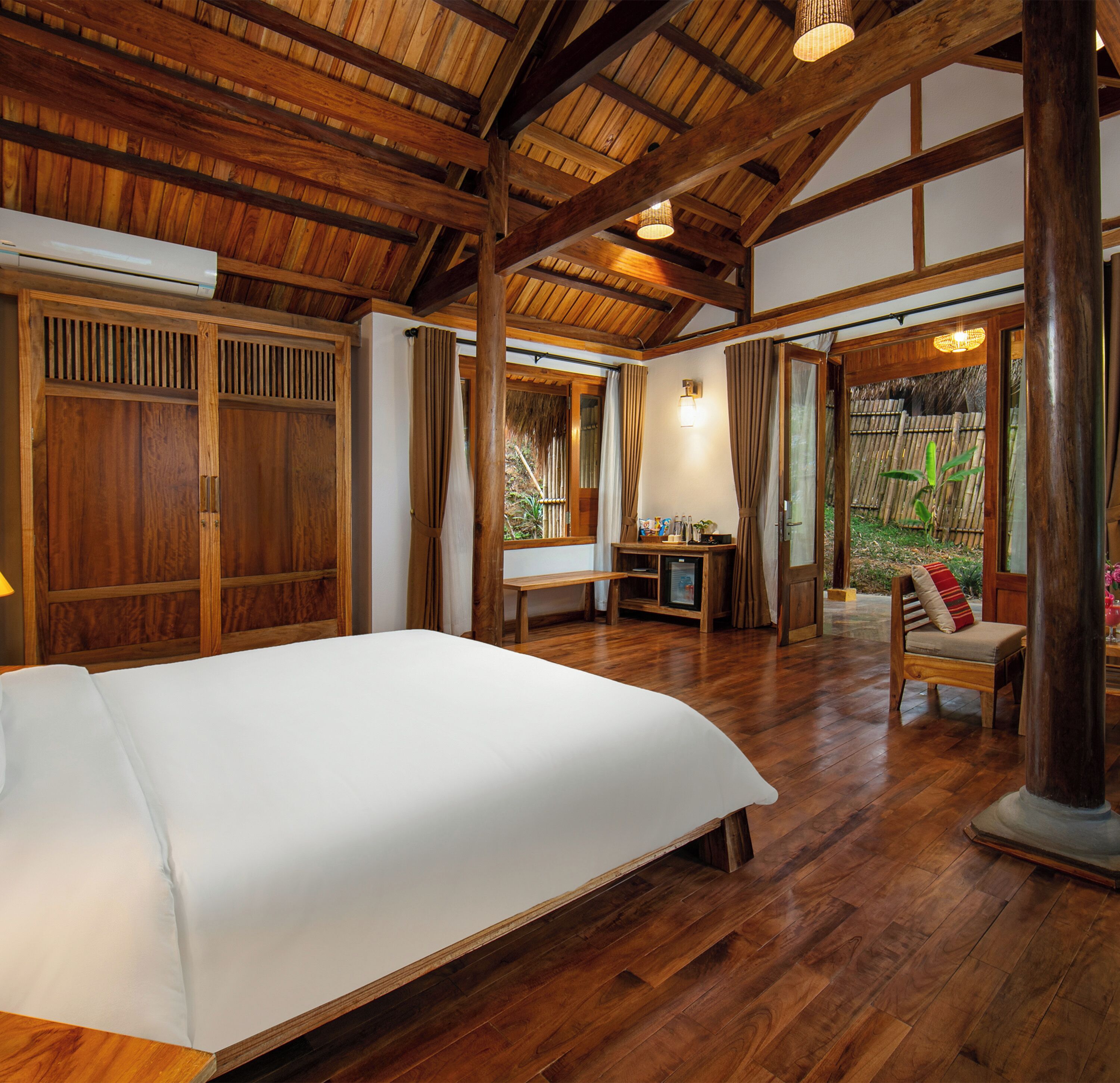 A spacious room with a large bed, wooden flooring, and a high ceiling with exposed beams. There is a small table with a chair and a window with curtains.