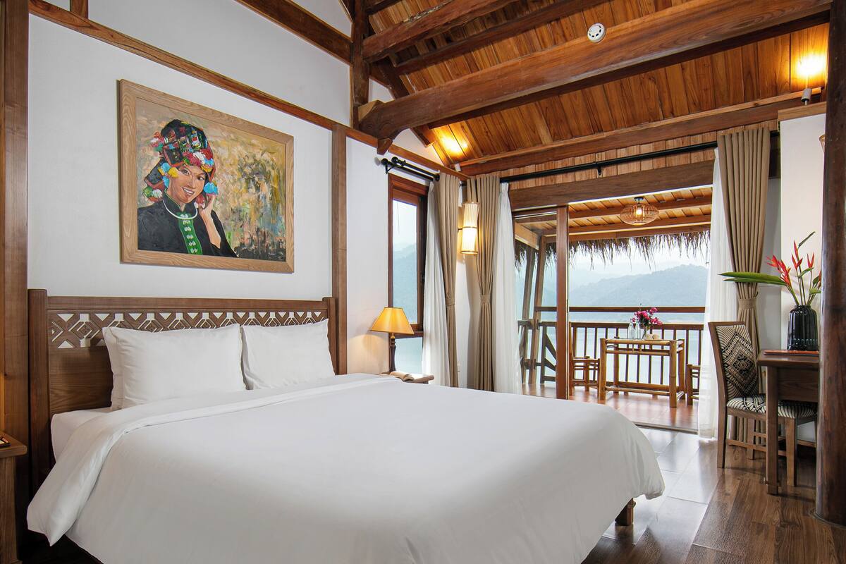 Une chambre spacieuse avec un grand lit, des poutres apparentes en bois au plafond, un tableau au mur et un balcon avec vue.
