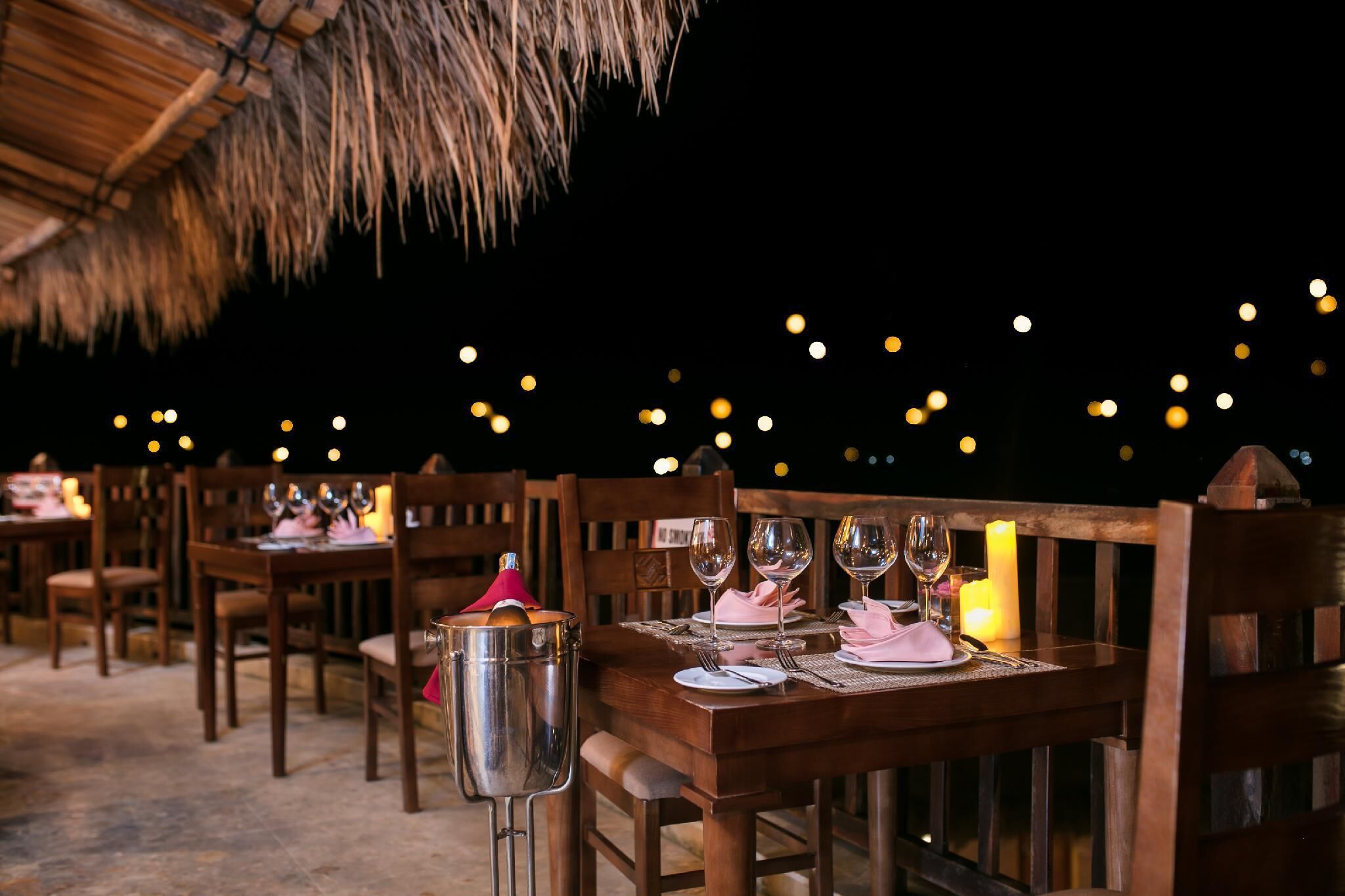A dining area with wooden tables and chairs, candles, and a thatched roof.