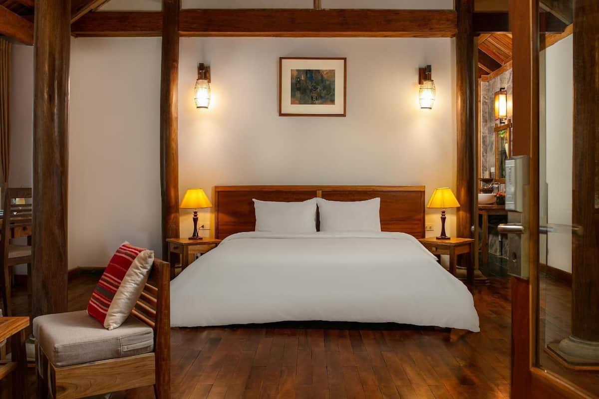 Une chambre à coucher bien aménagée, avec un grand lit, deux lampes de chevet, une tête de lit en bois et une seule chaise avec un coussin.