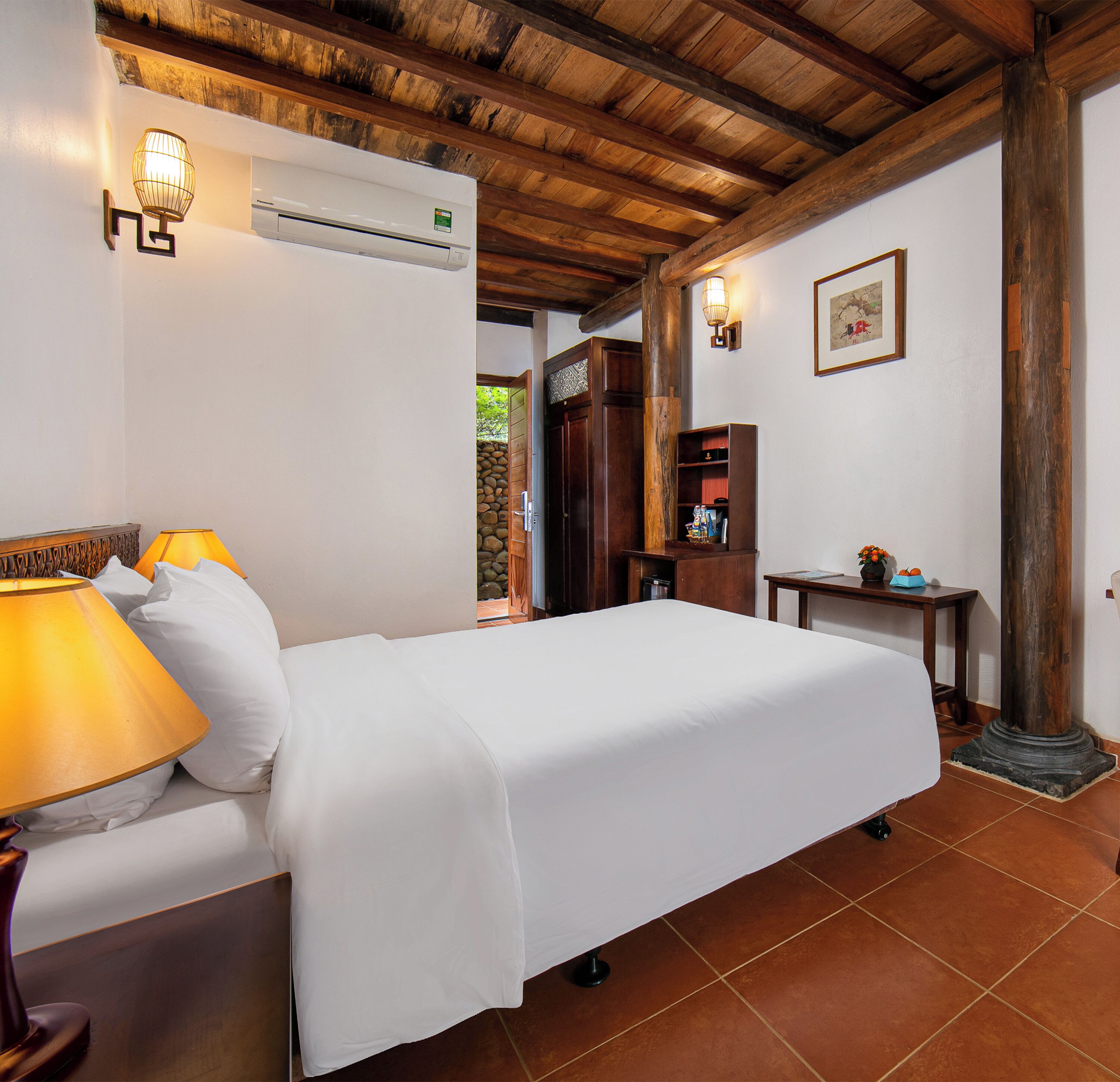 A hotel room with a bed, a lamp, a wall-mounted air conditioner, a wooden ceiling, and a small table with a vase.