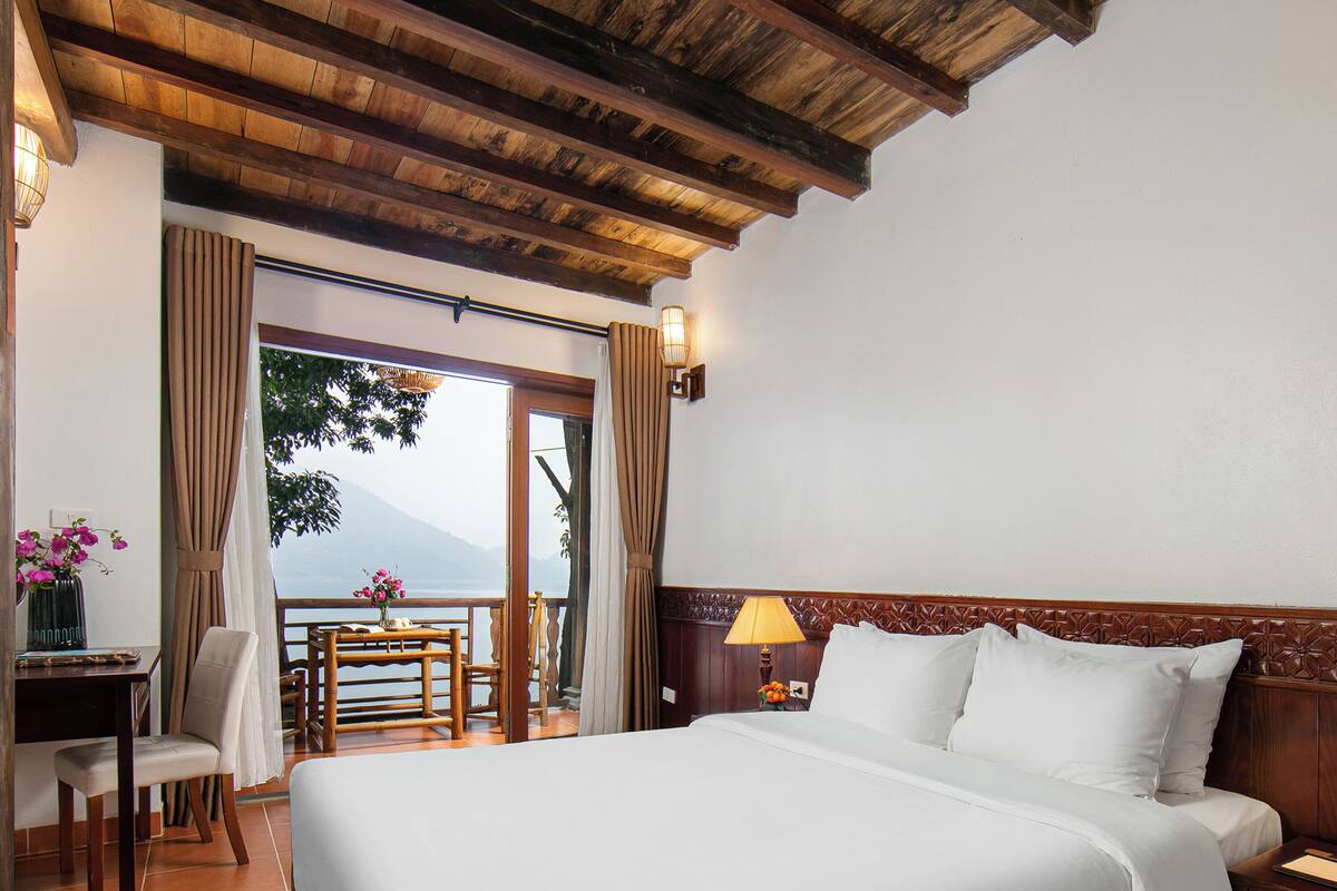 Une chambre d’hôtel avec un grand lit, un bureau avec une chaise, une lampe et une vue sur un lac et des montagnes, visible à travers une porte vitrée.