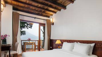 A hotel room with a large bed, a desk with a chair, a lamp, and a view of a lake and mountains through a glass door.