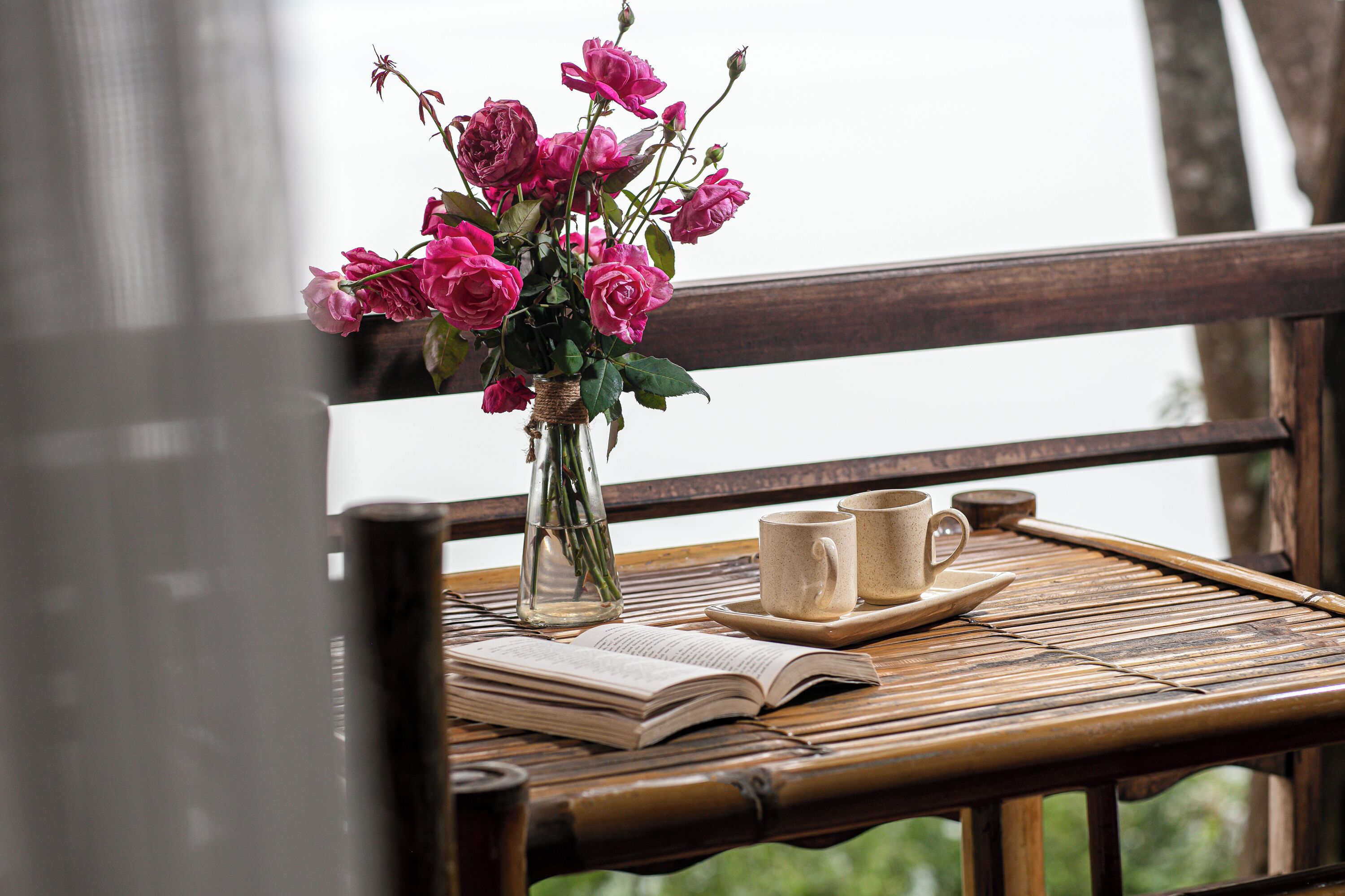 A wooden table with a book, a vase of pink flowers, and two coffee cups.