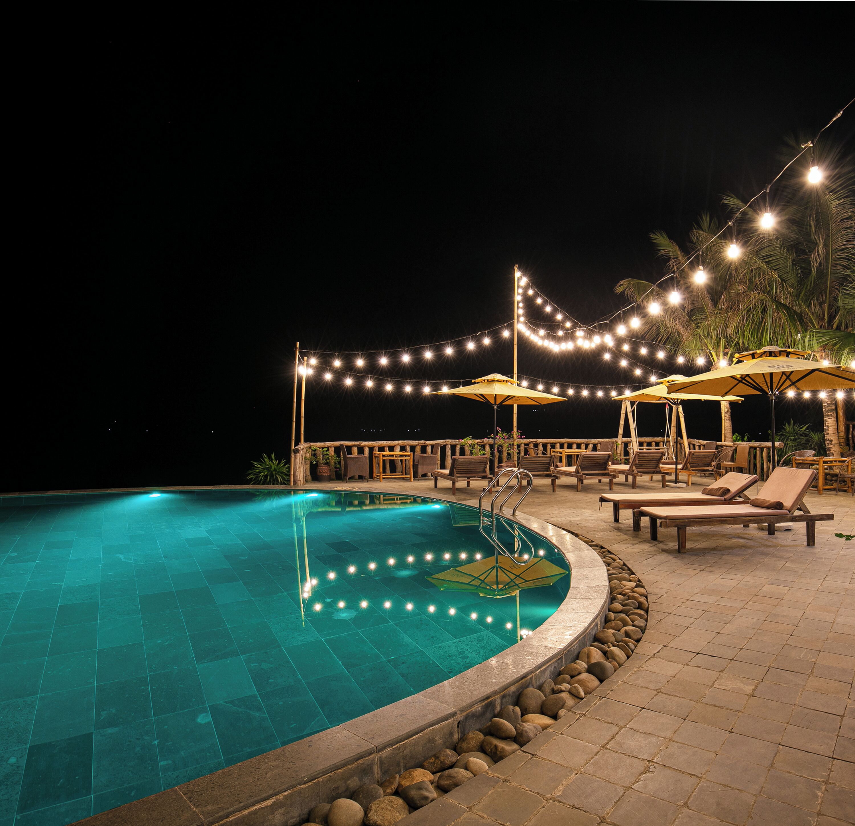 Una piscina nocturna con luces colgantes, sillones de descanso y sombrillas.