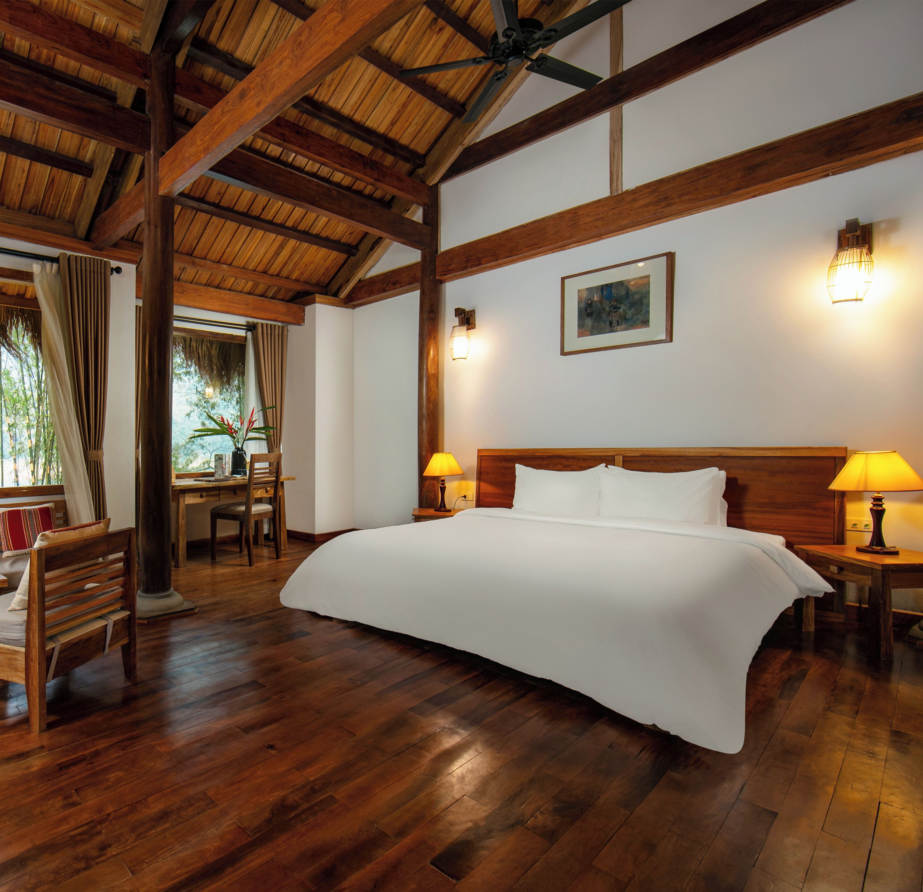 A spacious room with a large bed, wooden beams, a desk, and two lamps.