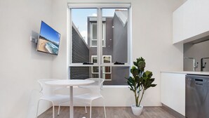 A modern kitchen with a dishwasher, a dining area with a white table and chairs, a large window with a view of a building, and a potted plant. - Bondi Beach Studios (Bondi)