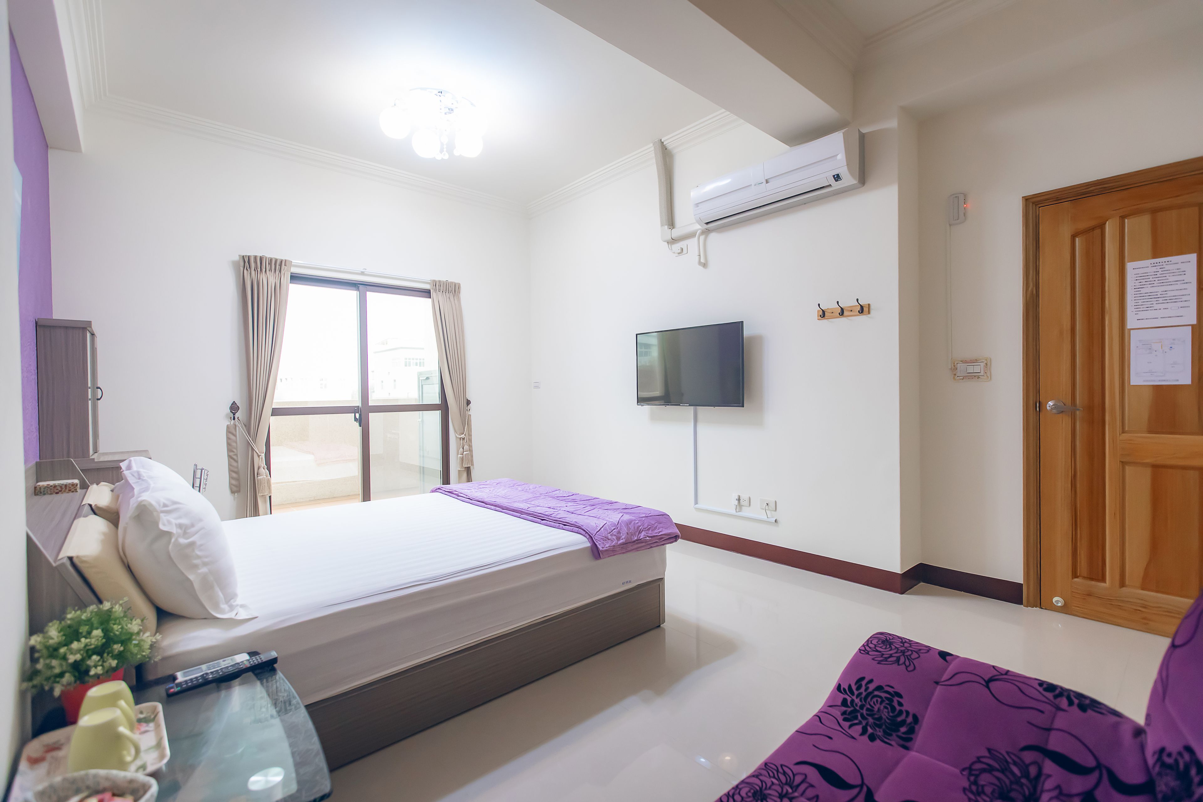 A hotel room with a bed, a TV, a window with curtains, an air conditioning unit, and a wooden door.