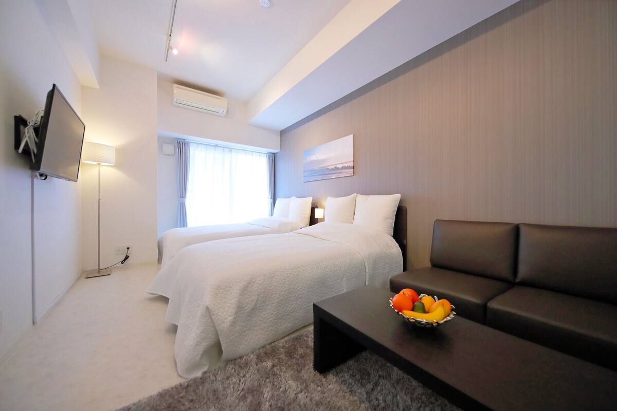 Une chambre d’hôtel moderne équipée d’un lit, d’un canapé, d’un téléviseur à écran plat et d’un panier de fruits posé sur une table basse.