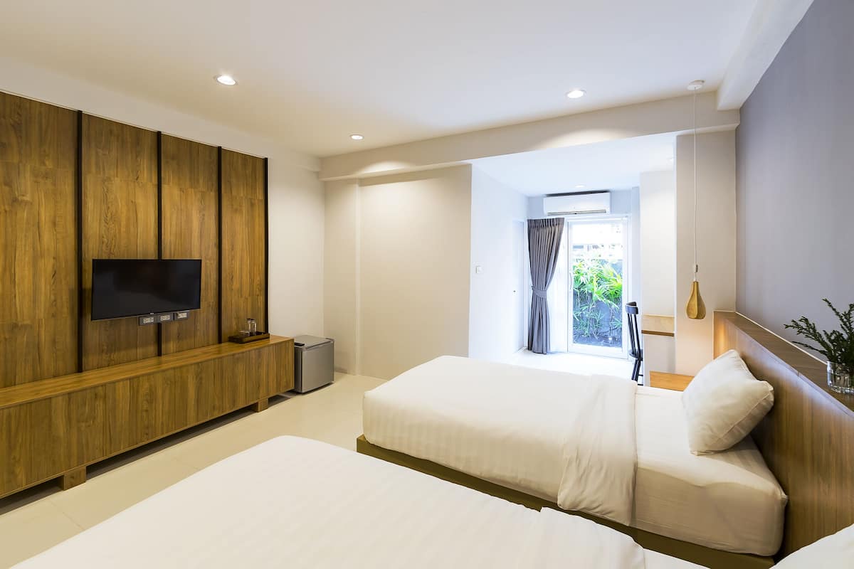 Habitación de hotel con dos camas, un televisor montado en un panel de madera, un cesto de basura pequeño y una ventana con cortinas.