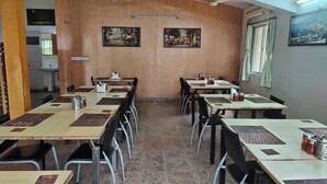 A dining area with tables and chairs, two wall-mounted paintings, and a window with curtains. -  Stay Guru Resort Farm Villa (Bhuj)
