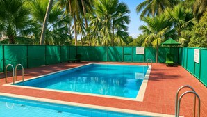 A rectangular swimming pool with clear blue water, surrounded by a red tiled deck and green fencing. -  Stay Guru Resort Farm Villa (Bhuj)