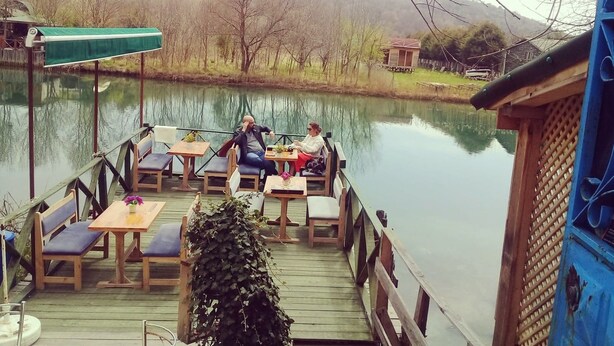 A wooden deck with tables and chairs overlooking a calm lake, surrounded by trees and a small building in the background. - Otel Turker (Sile)