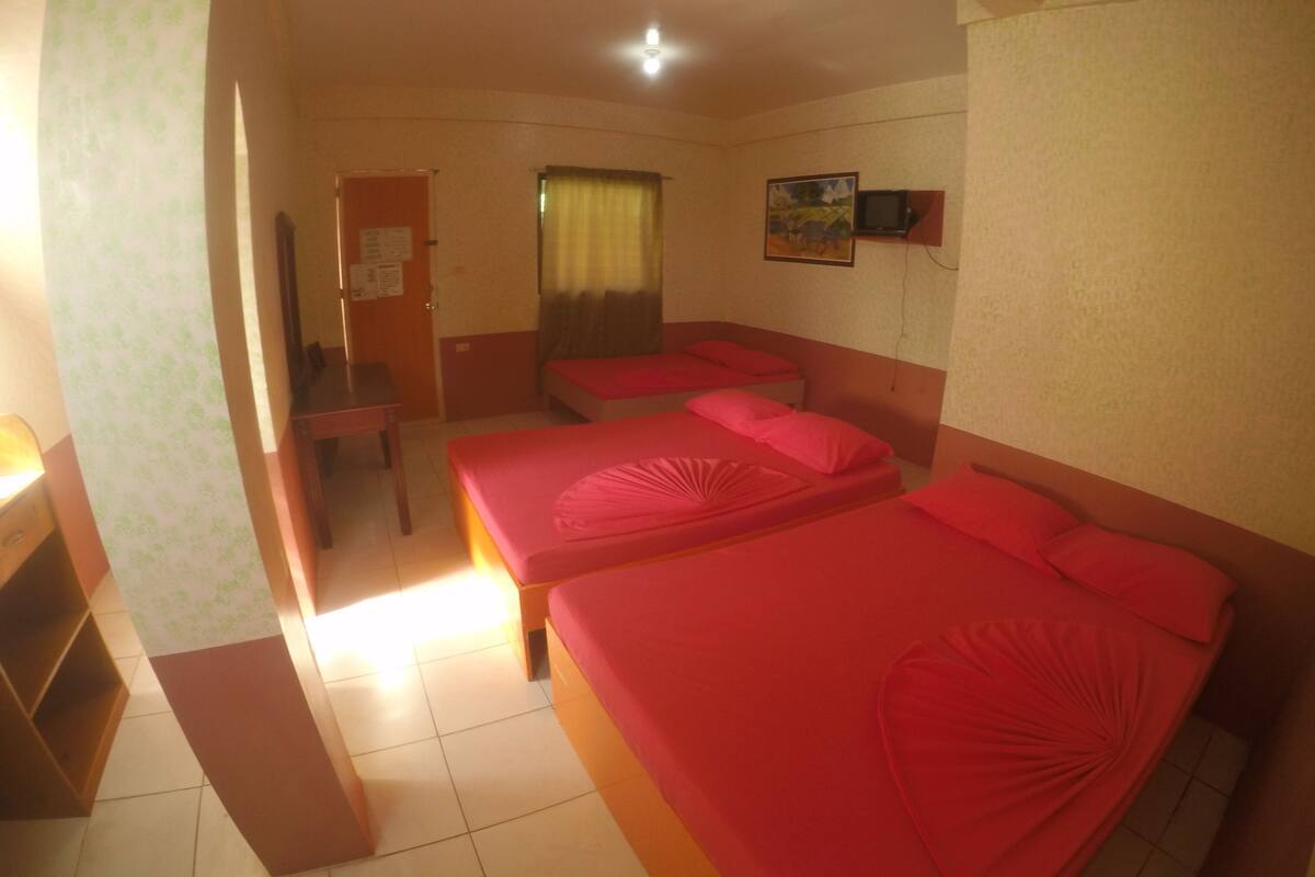 A hotel room with three beds, a desk, a chair, a TV, and a door with a notice.
