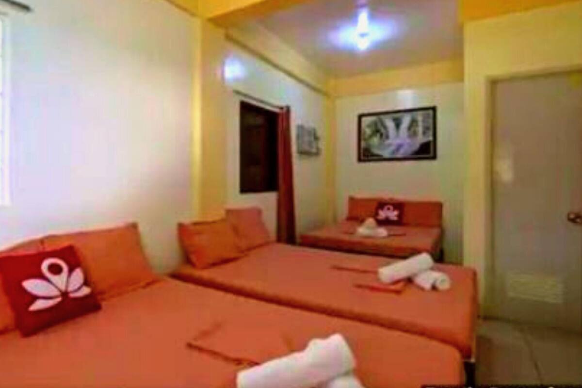 A room with two beds, each with orange bedding and pillows, a small table with a pillow, a window with blinds, and a door.