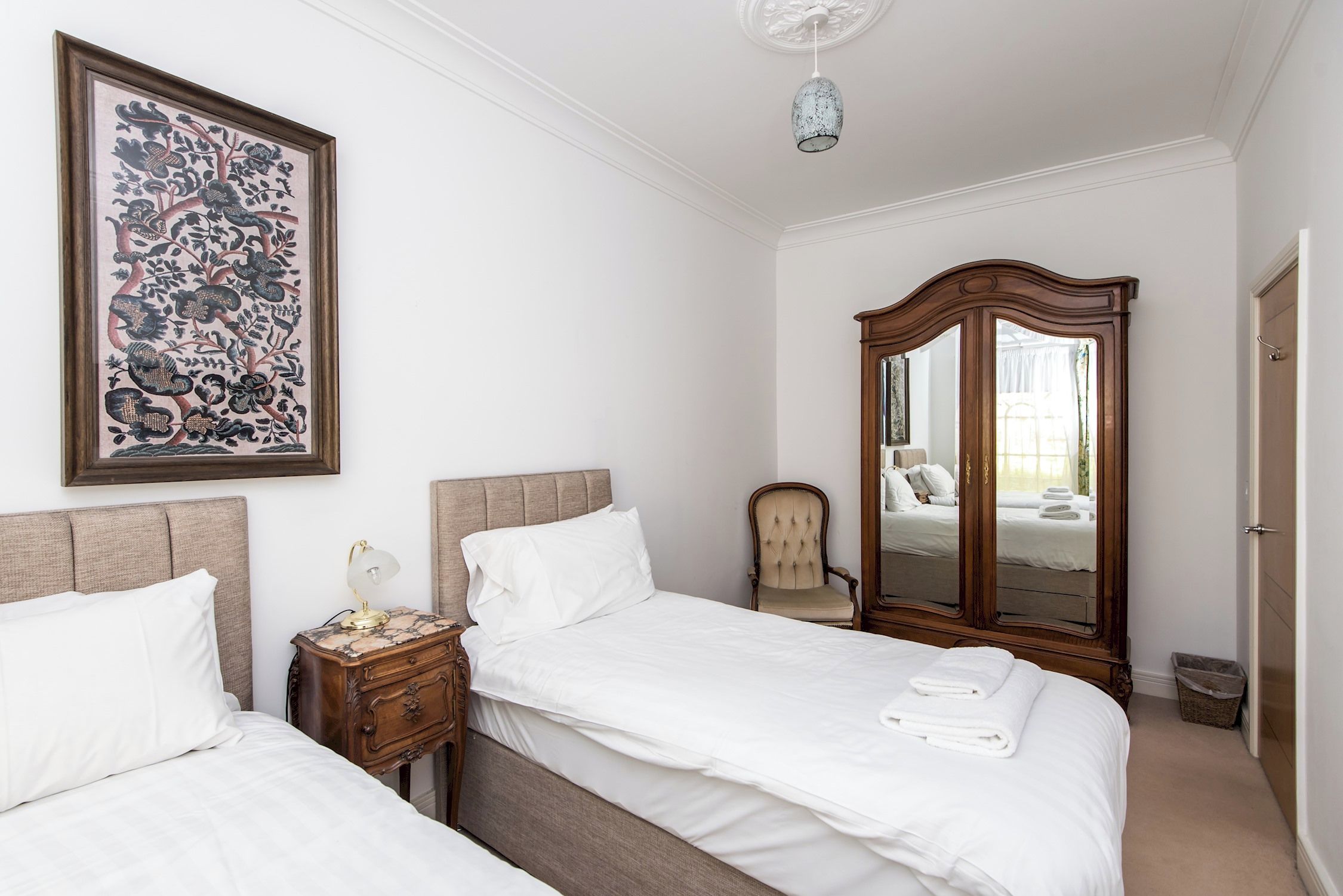 Quarto de hotel com duas camas, um espelho grande, uma mesa de cabeceira e uma obra de arte emoldurada na parede.