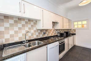 A modern kitchen with white cabinets, a black countertop, stainless steel appliances, and a checkered tile backsplash. - Toast Stays - The Three Rosellas at The Grove (Durham)