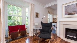 Una habitación con chimenea, piso de madera, una butaca tapizada de rojo, un sillón negro y vistas al exterior a través de amplias ventanas.