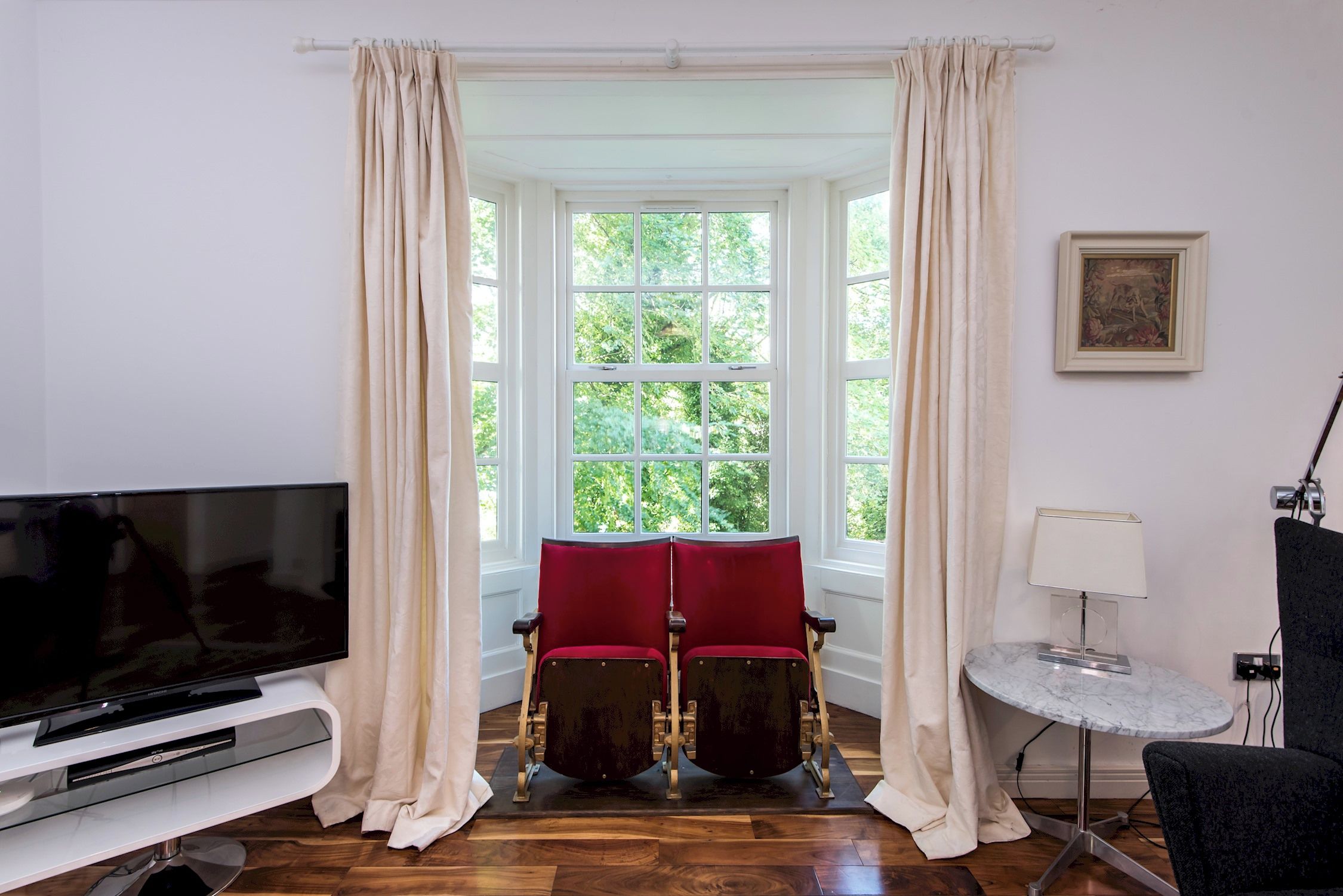 Une pièce avec une grande fenêtre, deux fauteuils rouges, un téléviseur et une petite table.