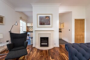 A living room with a fireplace, a dark armchair, a blue sofa, and a kitchen in the background.