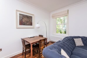 A room with a dining table, chairs, a framed picture, a floor lamp, a window with curtains, and a blue sofa. - Toast Stays - The Three Rosellas at The Grove (Durham)