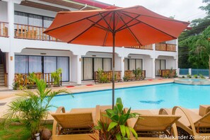 A poolside area with lounge chairs and an umbrella, surrounded by a building with balconies and windows. - Alona Vida Beach Hill (Panglao)