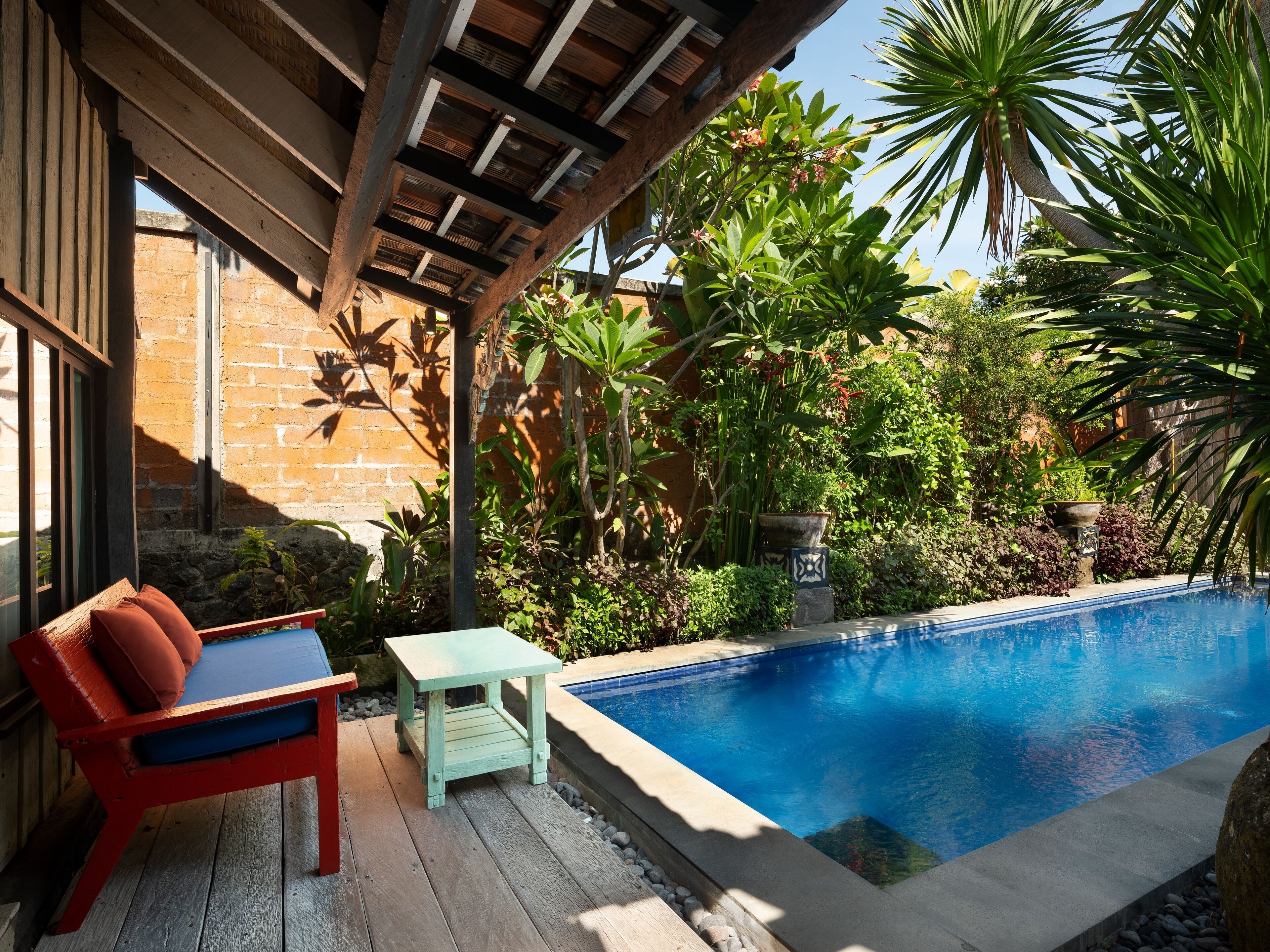 Zona junto a la piscina con cubierta de madera, una silla reclinable roja y azul, y una pequeña mesa.