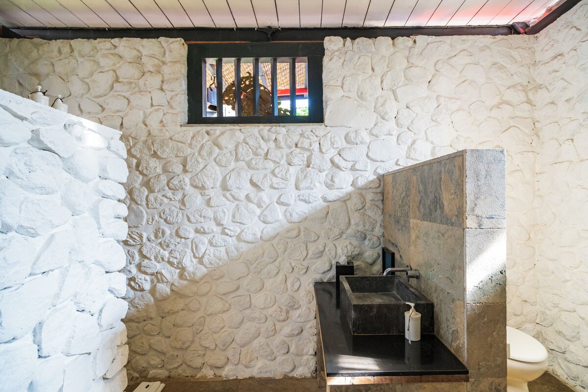 Une salle de bain avec un mur en pierre, une fenêtre grillagée, un lavabo et des toilettes.