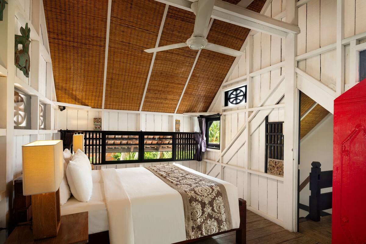 A room with a bed, a ceiling fan, and a view of greenery outside.