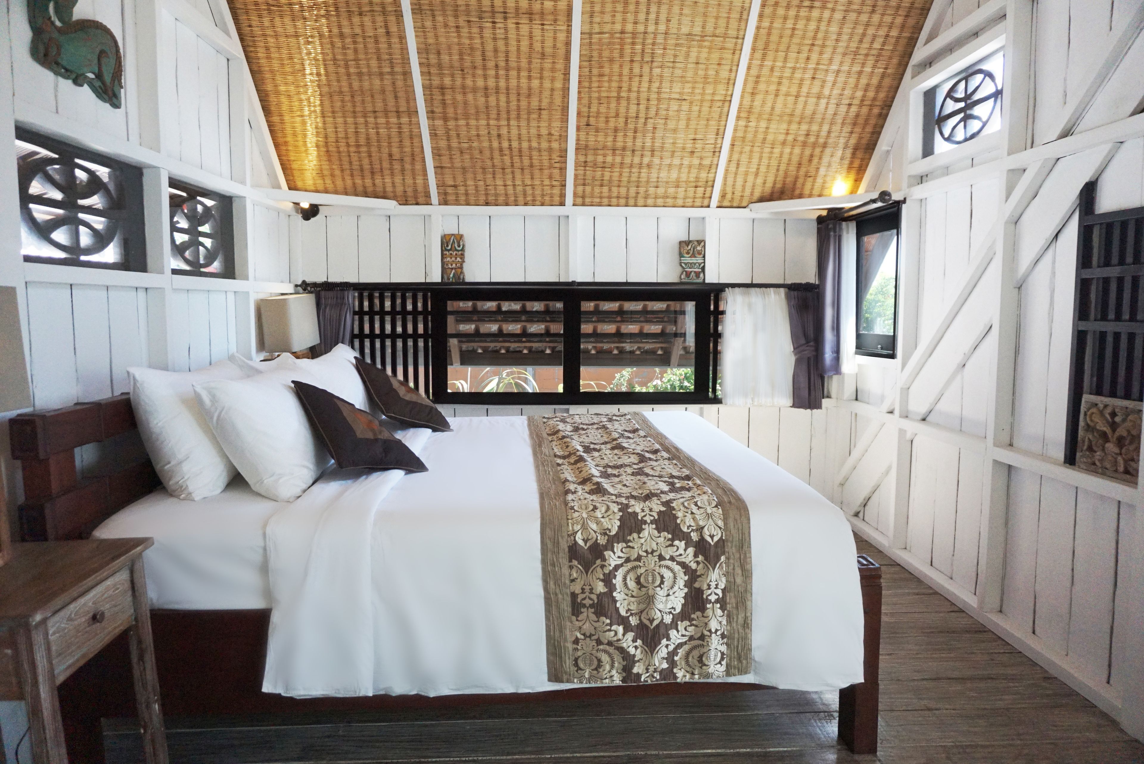 Una cama grande con colcha blanca y marrón, cabecero de madera y varios almohadones. Se ve una ventana con cortinas y vistas al exterior.