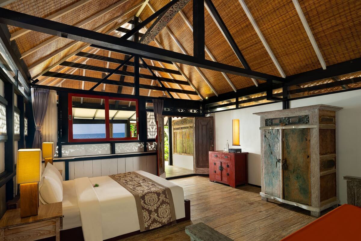 A spacious room with a high ceiling, a bed with a patterned bedspread, a wooden cabinet, and a view of the sea through a window.