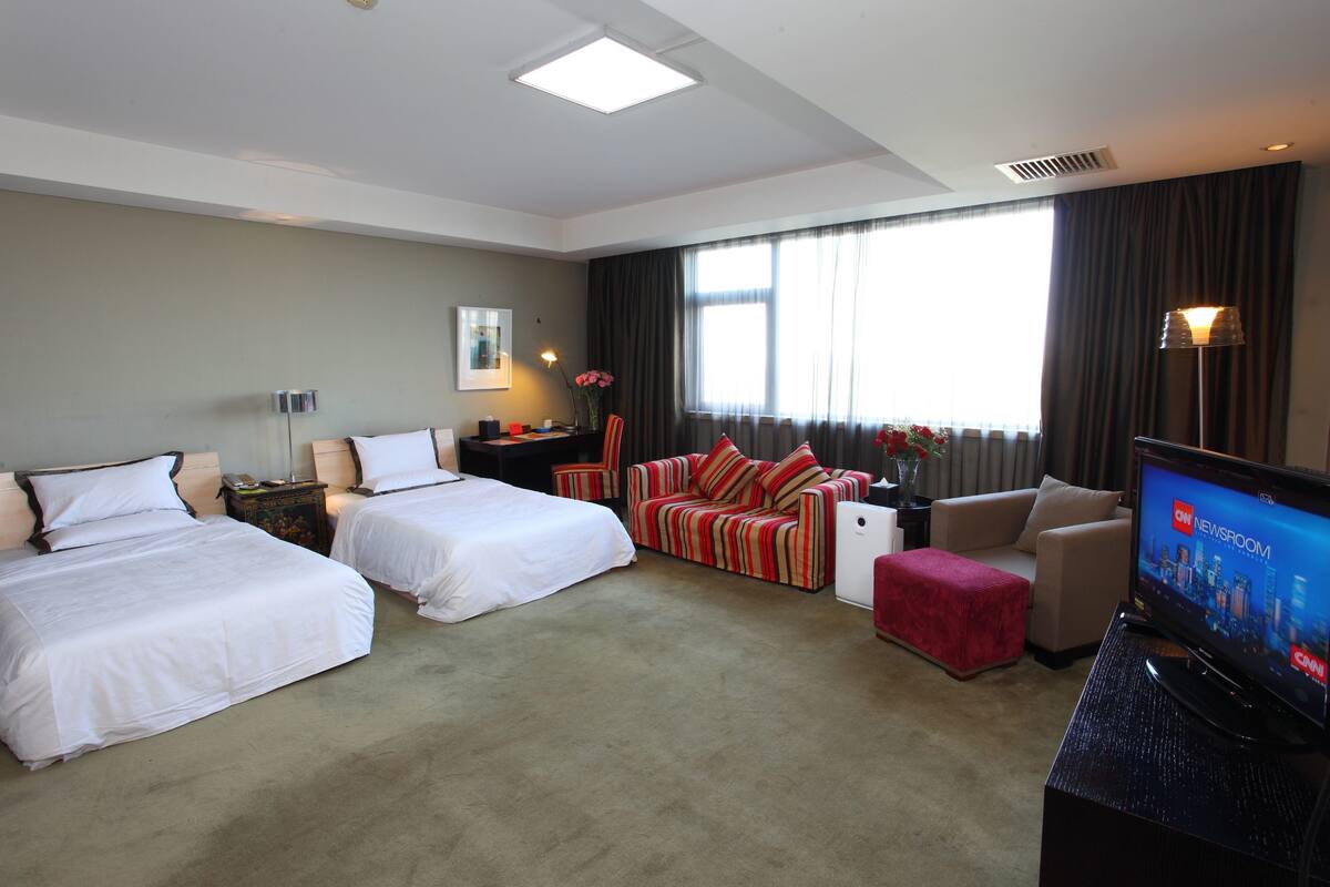 Quarto de hotel com duas camas, um sofá, uma televisão e uma escrivaninha com luminária.