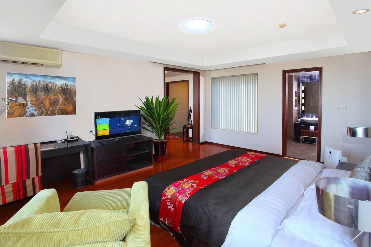 Quarto de hotel com uma cama grande, uma escrivaninha com computador, uma televisão, uma planta e um banheiro visível por uma porta aberta.